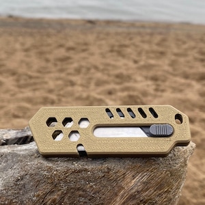 May include: A tan utility knife with a sliding blade and a textured surface. The knife has hexagonal and rectangular cutouts, and a small hole for a lanyard. The blade is silver, and the slider is grey.