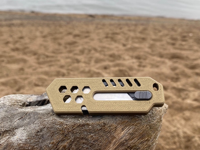 May include: A tan utility knife with a sliding blade and a textured surface. The knife has hexagonal and rectangular cutouts, and a small hole for a lanyard. The blade is silver, and the slider is grey.