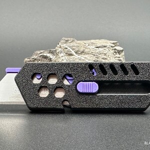 close up of a stylized, black utility knife with the blade extended in a ready-to-use position leaning against a rock against a white background. The knife has a purple button for the sliding mechanism and decorative holes and diagonal slats on the top.