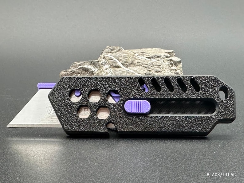 close up of a stylized, black utility knife with the blade extended in a ready-to-use position leaning against a rock against a white background. The knife has a purple button for the sliding mechanism and decorative holes and diagonal slats on the top.