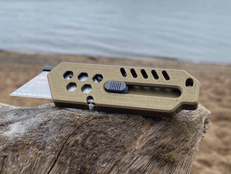 May include: A tan utility knife with a grey sliding mechanism and a silver blade. The knife has a textured design with hexagonal cutouts and is resting on a piece of driftwood.