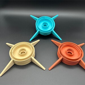 Three, different-colored, 3D-printed objects with a central circular design and four radiating arms. The central portion has concentric rings and a textured surface. The arms are flat and extend outward, creating a star-like appearance. One is tan, another is turquoise, and the third is terracotta colored.