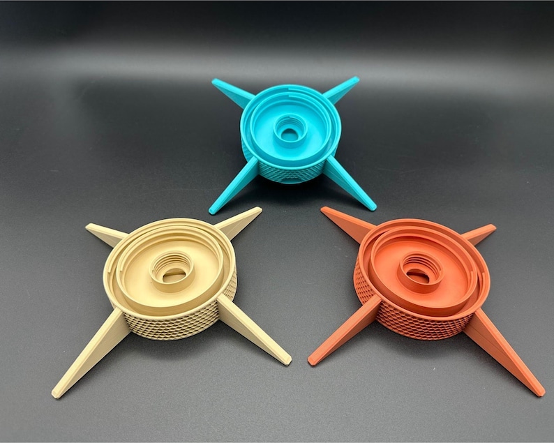 Three, different-colored, 3D-printed objects with a central circular design and four radiating arms. The central portion has concentric rings and a textured surface. The arms are flat and extend outward, creating a star-like appearance. One is tan, another is turquoise, and the third is terracotta colored.