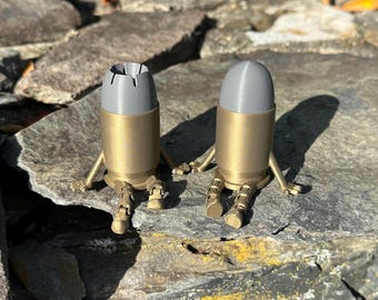 May include: Two novelty bullet-shaped figurines with brass-coloured bodies and grey tops. Each has small legs and arms. One has a split top, the other a rounded top. They are posed on a rocky surface.