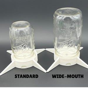 May include: Two clear glass Ball mason jars inverted on white plastic drying stands. The left stand is labeled STANDARD and the right stand is labeled WIDE-MOUTH. Each stand features a textured circular base with four pointed legs.