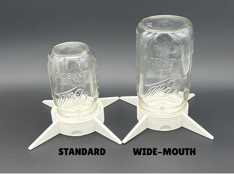 May include: Two clear glass Ball mason jars inverted on white plastic drying stands. The left stand is labeled STANDARD and the right stand is labeled WIDE-MOUTH. Each stand features a textured circular base with four pointed legs.