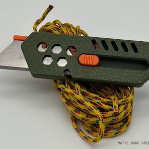 close up of a stylized, dark green utility knife with the blade extended in a ready-to-use position laying on top of a yellow-red-green patterned rope tidily coiled up all against a white background. The knife has an orange button for the sliding mechanism and decorative holes and diagonal slats on the top.