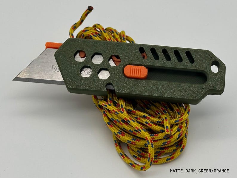 close up of a stylized, dark green utility knife with the blade extended in a ready-to-use position laying on top of a yellow-red-green patterned rope tidily coiled up all against a white background. The knife has an orange button for the sliding mechanism and decorative holes and diagonal slats on the top.