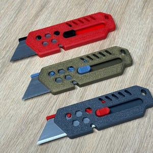 May include: Three utility knives in red, olive green, and dark grey. Each knife has a retractable blade and a coloured slider button. The handles have a textured surface and a series of holes and cutouts.