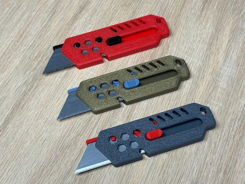May include: Three utility knives in red, olive green, and dark grey. Each knife has a retractable blade and a coloured slider button. The handles have a textured surface and a series of holes and cutouts.