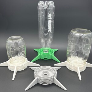 May include: Three bottle and jar drying stands with four-legged bases. Two white stands hold inverted glass jars, one green stand holds an inverted plastic bottle, and one grey stand sits empty. Each base features a textured, honeycomb-patterned rim.