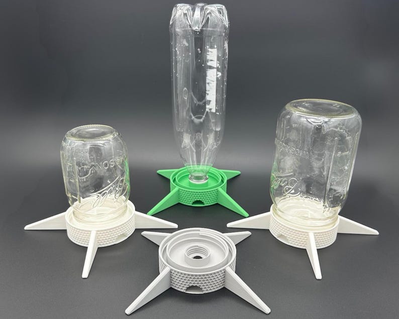 May include: Three bottle and jar drying stands with four-legged bases. Two white stands hold inverted glass jars, one green stand holds an inverted plastic bottle, and one grey stand sits empty. Each base features a textured, honeycomb-patterned rim.