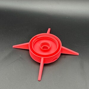 May include: A bright red, circular plastic component featuring a central threaded opening and four pointed, radiating arms extending outward. The exterior side wall of the central circular section displays a textured, cross-hatched grid pattern.