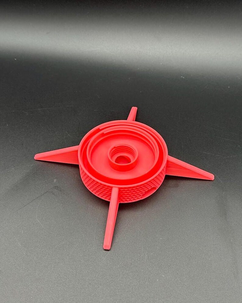 May include: A bright red, circular plastic component featuring a central threaded opening and four pointed, radiating arms extending outward. The exterior side wall of the central circular section displays a textured, cross-hatched grid pattern.