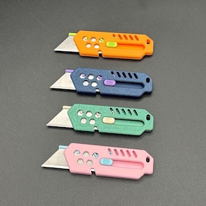 four, colored stylized utility knives with their blades extended and laying against a gray background. The knives are (top-bottom): orange, dark blue, metallic green, and baby pink. Each knife has decorative holes and diagonal slats on the top.