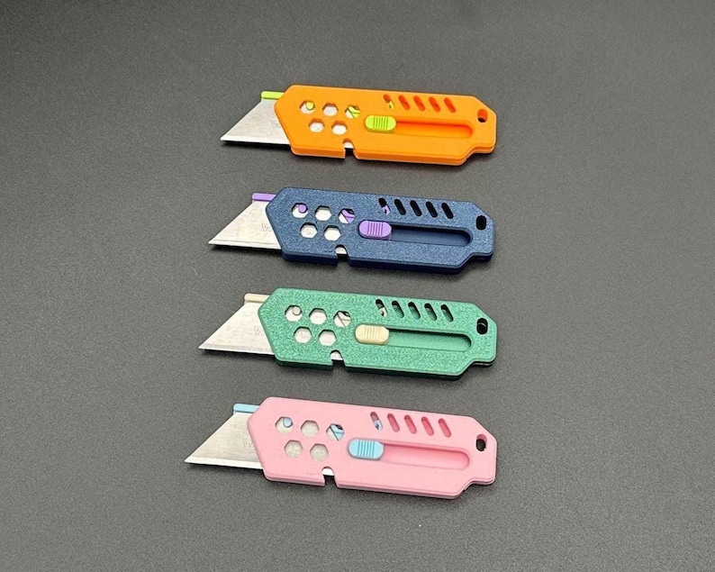 four, colored stylized utility knives with their blades extended and laying against a gray background. The knives are (top-bottom): orange, dark blue, metallic green, and baby pink. Each knife has decorative holes and diagonal slats on the top.