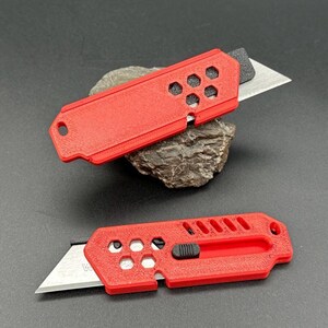 close up of two stylized, red utility knives with the blade extended in a ready-to-use position laying against a gray background. The knife has a black button for the sliding mechanism and decorative holes and diagonal slats on the top.