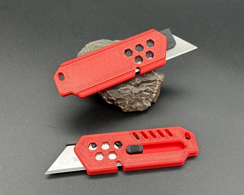 close up of two stylized, red utility knives with the blade extended in a ready-to-use position laying against a gray background. The knife has a black button for the sliding mechanism and decorative holes and diagonal slats on the top.