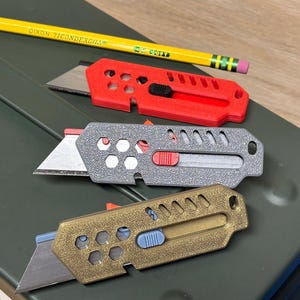 May include: Three utility knives in red, grey, and gold, with exposed blades and sliding mechanisms. A yellow pencil with the text "DIXON TICONDEROGA" rests above the knives. The knives are on a green surface.