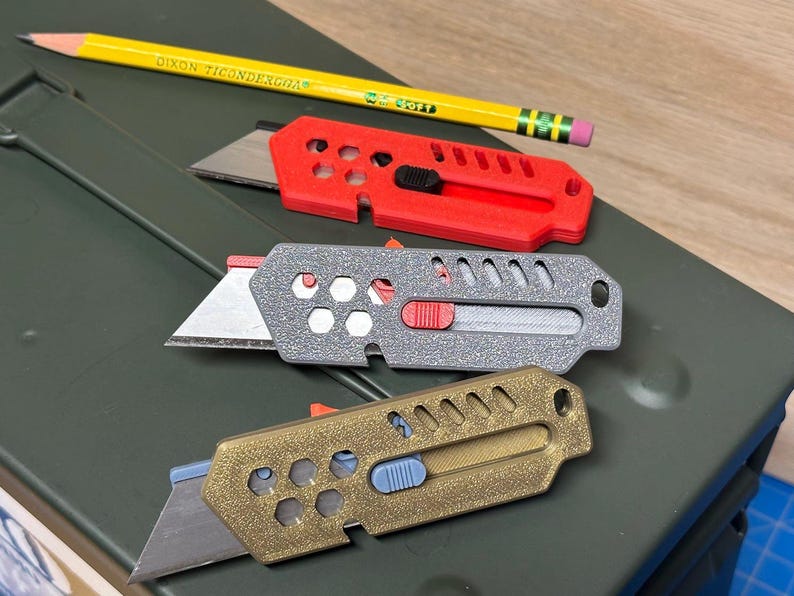 May include: Three utility knives in red, grey, and gold, with exposed blades and sliding mechanisms. A yellow pencil with the text "DIXON TICONDEROGA" rests above the knives. The knives are on a green surface.