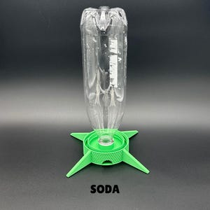 May include: A clear plastic bottle is inverted and attached to a bright green, four-legged circular stand with a textured, grid-like pattern on its side. The word SODA is printed in black capital letters below the green stand on a dark background.