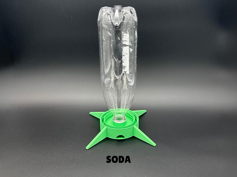 May include: A clear plastic bottle is inverted and attached to a bright green, four-legged circular stand with a textured, grid-like pattern on its side. The word SODA is printed in black capital letters below the green stand on a dark background.