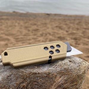 May include: A tan utility knife with a hexagonal pattern on the end and a silver blade. The knife is resting on a piece of driftwood, with a blurred background of a beach and sea.