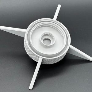May include: A light gray plastic component featuring a central threaded circular opening, a raised inner ring, and four long, pointed arms extending outward from a textured, diamond-patterned cylindrical base. The object is rigid and symmetrical.