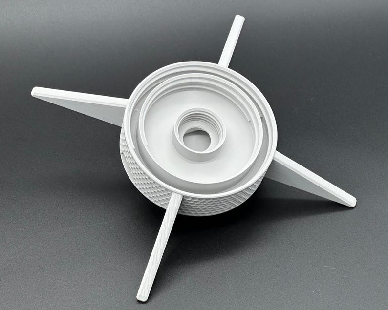 May include: A light gray plastic component featuring a central threaded circular opening, a raised inner ring, and four long, pointed arms extending outward from a textured, diamond-patterned cylindrical base. The object is rigid and symmetrical.