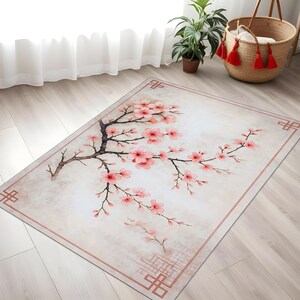 Op de afbeelding: Rechthoekig vloerkleed met een aquarelontwerp van een kersenbloesemtak met roze bloemen en bruine takken. Het vloerkleed heeft een lichtbeige achtergrond met een roze rand en decoratieve hoekelementen.