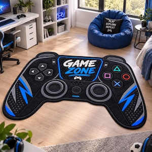 Puede incluir: Una alfombra negra con forma de mando de videojuego con detalles en azul y gris. El centro presenta el texto "GAME ZONE" en letras blancas y azules, rodeado de botones direccionales, joysticks circulares y símbolos geométricos.