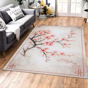 Op de afbeelding: Rechthoekig vloerkleed met een aquarelontwerp van een kersenbloesemtak met roze bloemen en bruine takken. Het vloerkleed heeft een lichtbeige achtergrond en een roze rand met hoekontwerpen.
