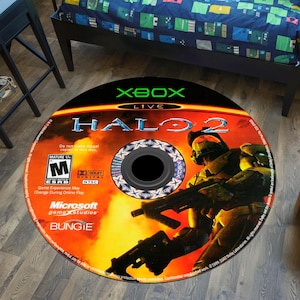 May include: Round rug featuring the Halo 2 video game disc design. The rug has a black center with a detailed image of a soldier holding a weapon, set against a fiery background. The Xbox Live and Halo 2 logos are prominently displayed.