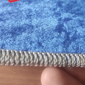 Op de afbeelding: Een close-up van een blauw fluwelen tapijt met een zilveren gevlochten rand. Het tapijt heeft een textuur en een "SAMPLE" stempel in rood.