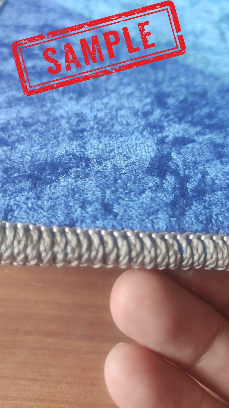 Op de afbeelding: Een close-up van een blauw fluwelen tapijt met een zilveren gevlochten rand. Het tapijt heeft een textuur en een "SAMPLE" stempel in rood.