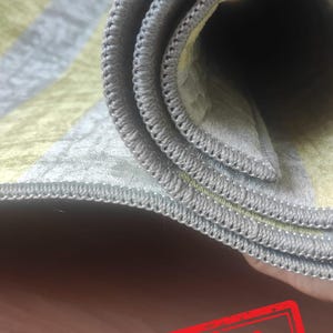 May include: A rolled area rug featuring alternating stripes of soft yellow and light gray with a subtle textured pattern. The edges of the rug are finished with a tight, gray overlock stitch. A red rectangular stamp with the text SAMPLE is visible.