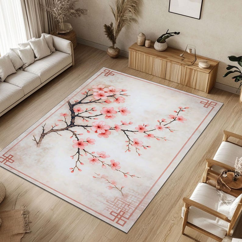 Op de afbeelding: Rechthoekig vloerkleed met een kersenbloesem ontwerp. Het kleed heeft een lichtbeige achtergrond, een roze rand en een tak met roze bloemen. Het kleed ligt op een houten vloer in een woonkamer.