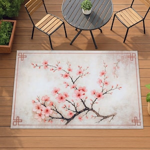 Op de afbeelding: Rechthoekig buitenkleed met een aquarelontwerp van roze kersenbloesems op een tak. Het kleed heeft een lichtbeige achtergrond met een roze rand en decoratieve hoekelementen. Het kleed ligt op een houten terras.