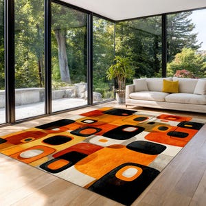 Op de afbeelding: Een rechthoekig vloerkleed met een modern design uit het midden van de eeuw. Het vloerkleed heeft een patroon van oranje, zwarte en crèmekleurige geometrische vormen. Het vloerkleed ligt op een houten vloer in een kamer met grote ramen.
