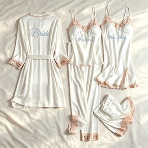Puede incluir: Un conjunto de ropa de dormir de satén blanco que incluye una bata con la palabra Bride, un conjunto de camisola y pantalón con Mrs. Aria, y una camisola con shorts. Cada pieza tiene detalles de encaje rosa claro en los bordes y mangas.