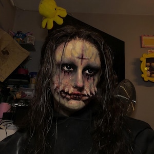 Peut inclure: Une personne portant un masque prothétique au teint pâle et texturé, avec une croix sombre sur le front et une large bouche ensanglantée aux dents apparentes. Le masque présente des orbites sombres et de longs cheveux ondulés, avec un sweat noir.