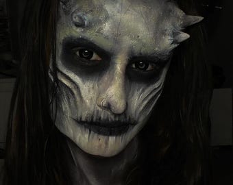 May include: A face featuring intricate prosthetic makeup with pale, textured skin, prominent forehead horns, and deep black shading around the eyes and mouth. The look includes dark, sunken features, a small nose ring, and long dark hair framing the face.