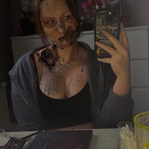 May include: A person with zombie makeup, featuring decaying skin and exposed wounds. They are wearing a black top and a gray zip-up hoodie. The makeup includes dark colors and blood effects, creating a gruesome appearance.