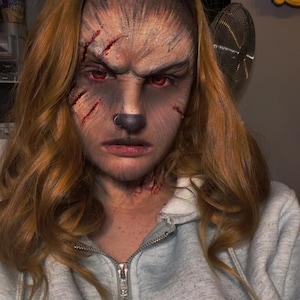 May include: A person with long wavy auburn hair wearing a light grey zip-up hoodie. The face features detailed werewolf-style makeup with red eyes, a dark nose, and simulated facial scars with red colouring on the forehead and cheek area.