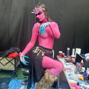 Op de afbeelding: Een persoon met roze lichaamsverf en blauwe vingernagels, gekleed in een zwarte rok met botaccenten en een harige beenwarmer. De persoon heeft een gehoornde hoofdtooi en zit in de buurt van een tafel met make-up benodigdheden.