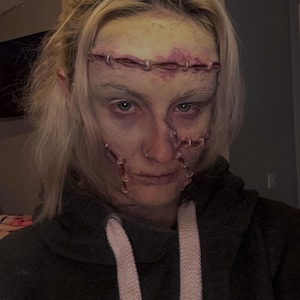 May include: A person with blonde hair is wearing a dark grey hoodie with a white drawstring and a white logo that says "LEICESTER UNIVERSITY DE MONCTON IT". The person has makeup on their face that makes them look like a monster with stitches.