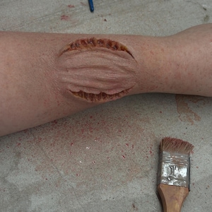 May include: A close-up of a fake wound on a person's arm. The wound is made of a brown material and has a jagged edge. A paintbrush is on the surface next to the arm.