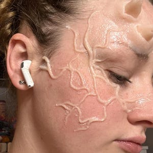 Può includere: Primo piano del viso di una persona con un auricolare bianco nell'orecchio. La pelle è adornata da una rete di strutture chiare, simili a vene, forse trucco o un effetto speciale. La pelle ha una tonalità leggermente rosata.