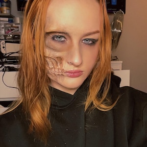 May include: A person with ginger hair and blue eyes, wearing a black hoodie with a "Toy Story" logo. The person has a half-skull makeup design on their face, creating a spooky Halloween look.