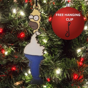 Może przedstawiać: Ozdoba świąteczna z Homerem Simpsonem. Ozdoba przedstawia Homera w jego klasycznym stroju, z białą koszulą, niebieskimi spodniami i żółtą głową. Ozdoba wisi na czarnym sznurku na tle choinki z czerwonymi i białymi światłami.